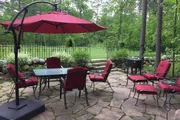 This image depicts a peaceful outdoor patio area with red cushioned chairs, a table under a red umbrella, and a grill, all set on flagstone paving with a lush green background of a well-kept garden and trees.