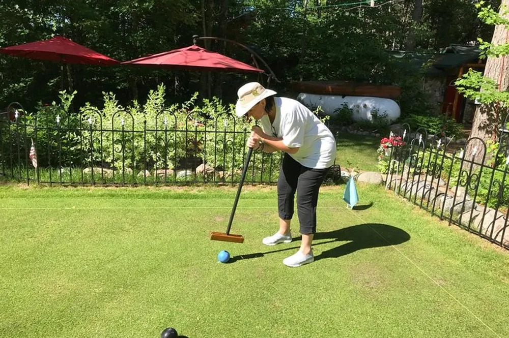 A person is playing croquet on a sunny day with a garden and red umbrellas in the background