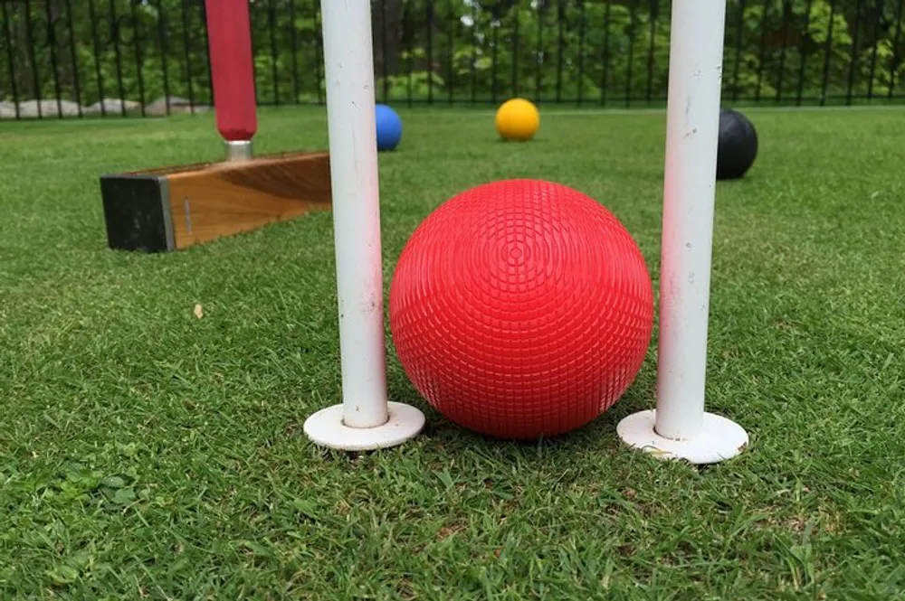 The image shows a red croquet ball positioned close to a hoop on a grassy lawn with other balls and a mallet in the background suggesting an ongoing game of croquet