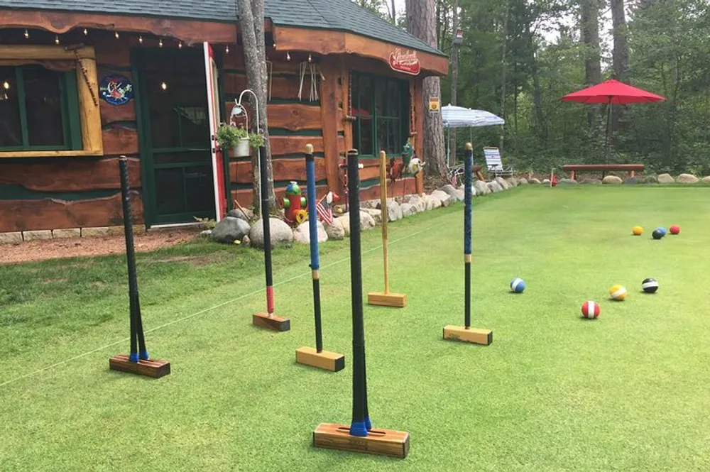 This image shows an outdoor croquet setup on green grass with colorful balls scattered across the playing field and a rustic wooden cabin in the background