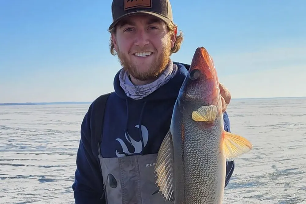 A smiling person wearing a cap and outdoor gear is holding a large fish with a snowy icy landscape in the background