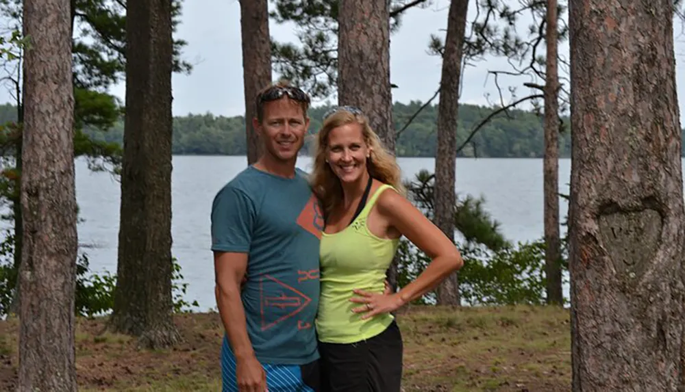 A man and a woman are smiling and posing for a photo between the trunks of pine trees with a serene lake in the background