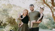 A smiling family of four stands together outdoors, with two adults each holding a young child, surrounded by greenery.