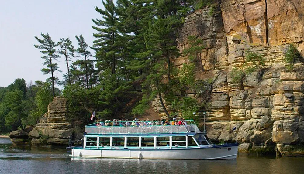 A tour boat filled with passengers is seen cruising near a steep cliff adorned with green trees