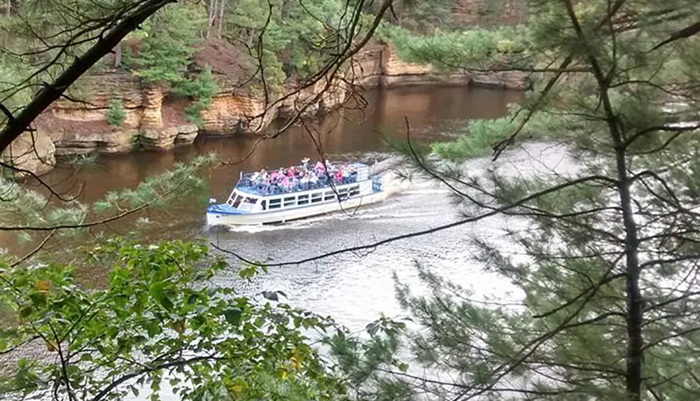 A tour boat is carrying passengers along a river bordered by forested cliffs viewed through a frame of leafy branches