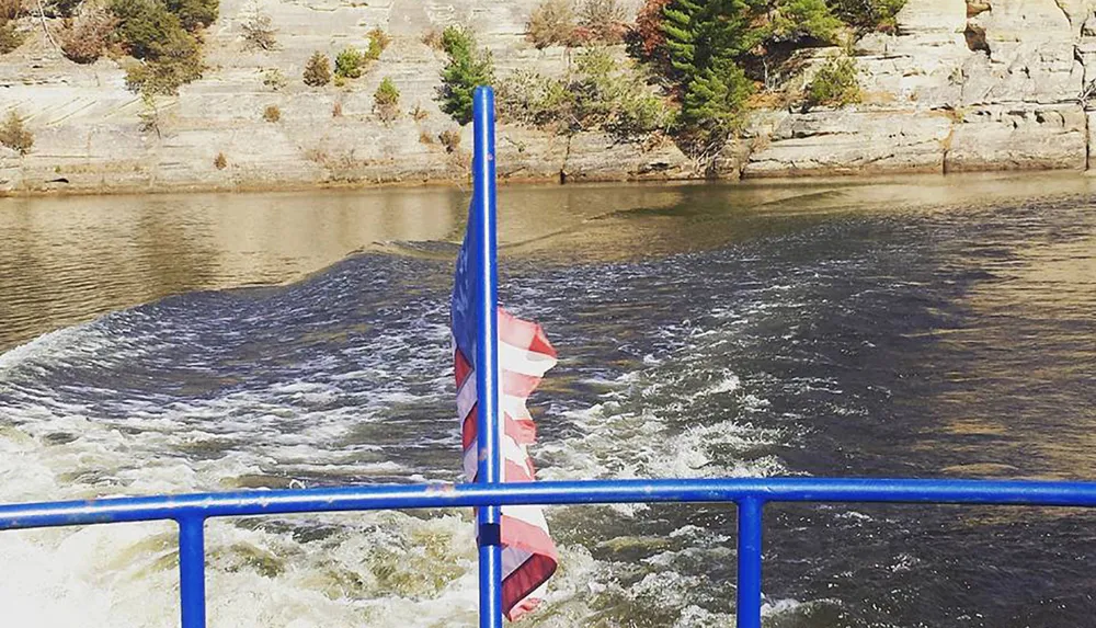 The image shows a rippled body of water in front of a rock face with the United States flag partly visible on a pole all framed by a blue metal railing