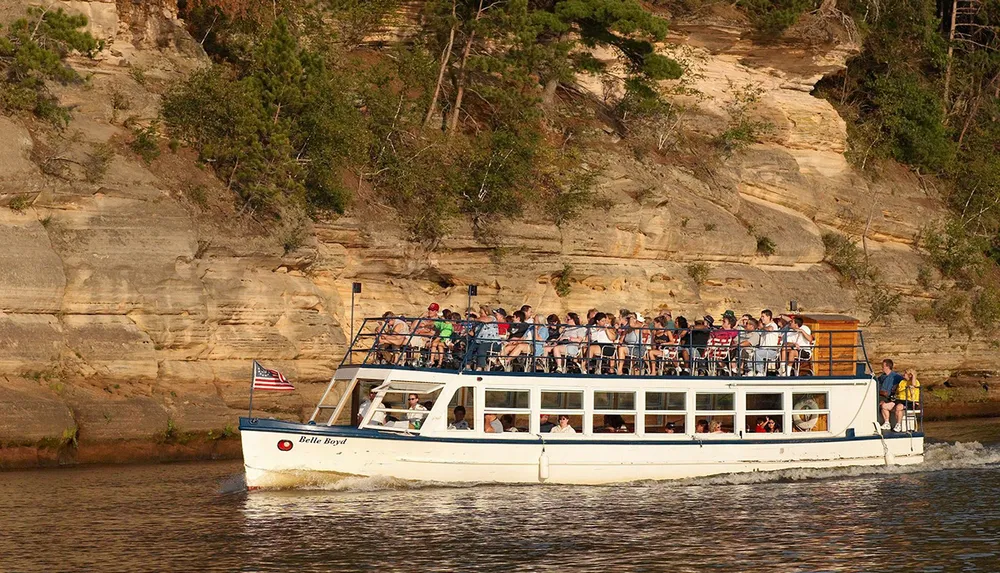 A packed tour boat named Belle Boyd is cruising near a sandstone cliff with passengers enjoying the view