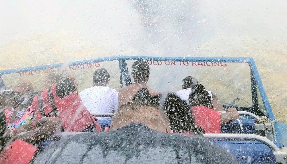 Passengers in life jackets are getting soaked while on a boat ride near a waterfall as evidenced by the spray and the HOLD ON TO RAILING sign
