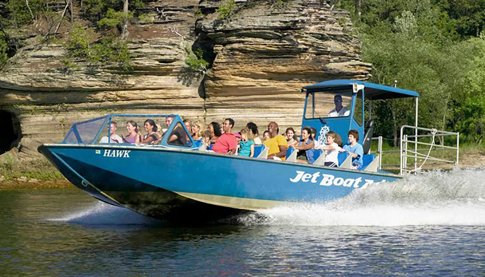 A group of people are enjoying a ride on a blue jet boat named HAWK on a sunny day with natural rock formations in the background