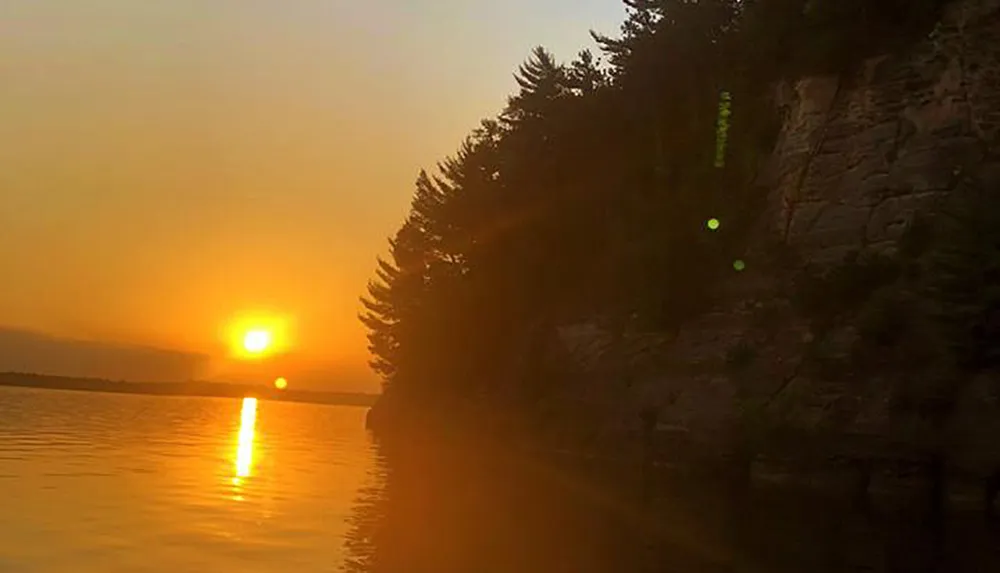 The image captures a golden sunset over a tranquil lake with the suns reflection on the water and a silhouette of a forested cliff to the right