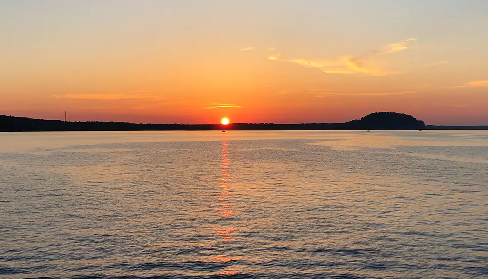 The image captures a tranquil sunset over a body of water with the sun close to the horizon casting an orange glow in the sky and reflecting on the waters surface