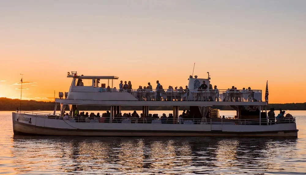 A multi-deck tour boat is filled with passengers enjoying a scenic sunset cruise on calm waters