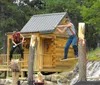 Two individuals wearing plaid shirts are engaging in a log sawing competition outside a shack with various tools and saws laid out around them