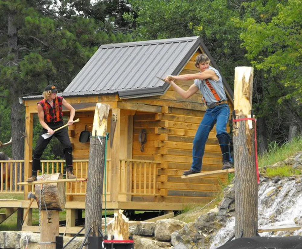Two individuals wearing outdoors or lumberjack attire are engaged in a log chopping demonstration with one standing on a horizontal log mid-swing with an axe and the other balancing on an upright log preparing to slice it with a saw