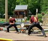 Two individuals wearing plaid shirts are engaging in a log sawing competition outside a shack with various tools and saws laid out around them