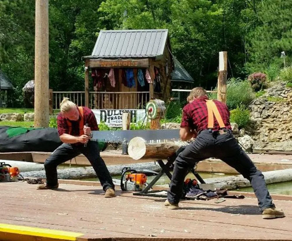 Two individuals wearing plaid shirts are engaging in a log sawing competition outside a shack with various tools and saws laid out around them