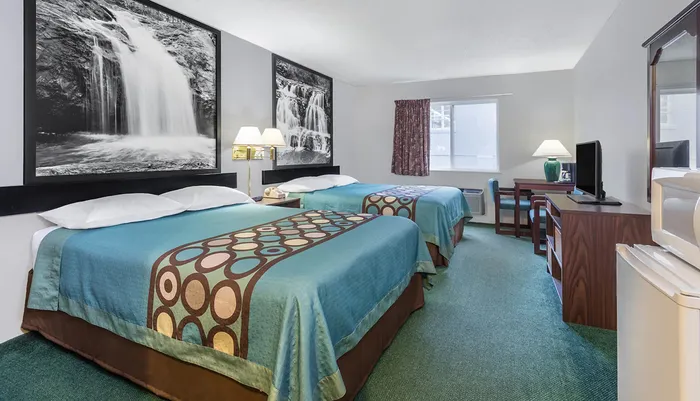 This image shows a neatly arranged hotel room with two beds patterned bedcovers a large waterfall photograph above the headboards a desk with a TV and a window with curtains