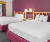 The image shows a neat hotel room with two queen-sized beds a rustic wood headboard white linens and a purple accent wall