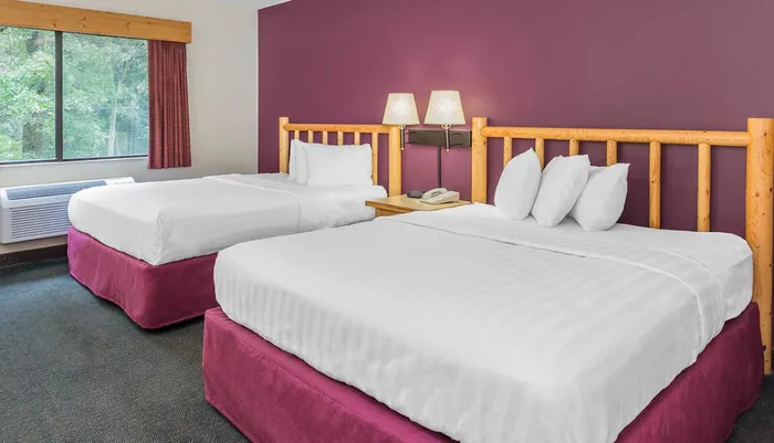 The image shows a neat hotel room with two queen-sized beds a rustic wood headboard white linens and a purple accent wall