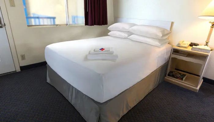 The image shows a neatly made single bed in a simple hotel room with a white and grey color scheme complemented by a nightstand and lamp on the side