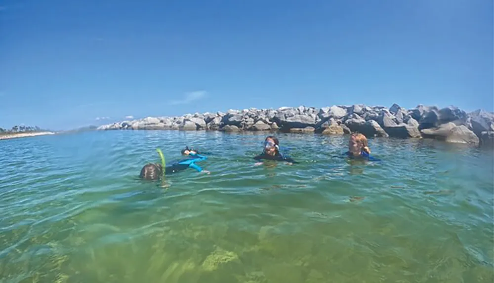 A group of people are enjoying a snorkeling activity in clear shallow waters near a rocky breakwater under a blue sky