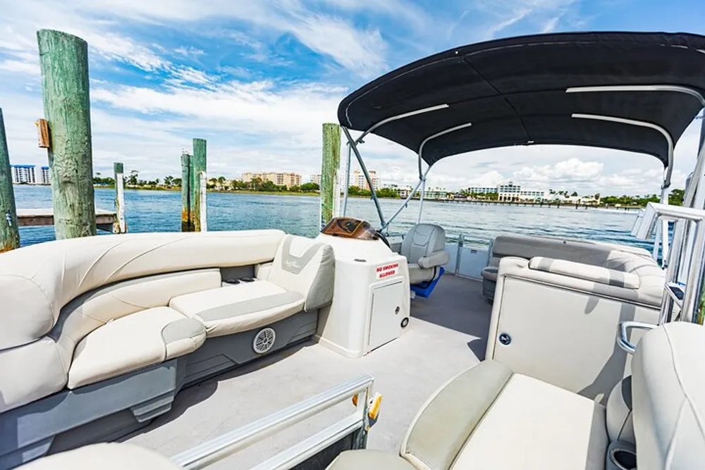 The image shows the interior of a pontoon boat with comfortable seating docked in a marina with a view of clear skies and coastal buildings in the background