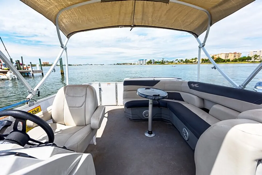 The image shows the interior of a pontoon boat with comfortable seating and a view of a calm body of water and waterfront buildings in the background