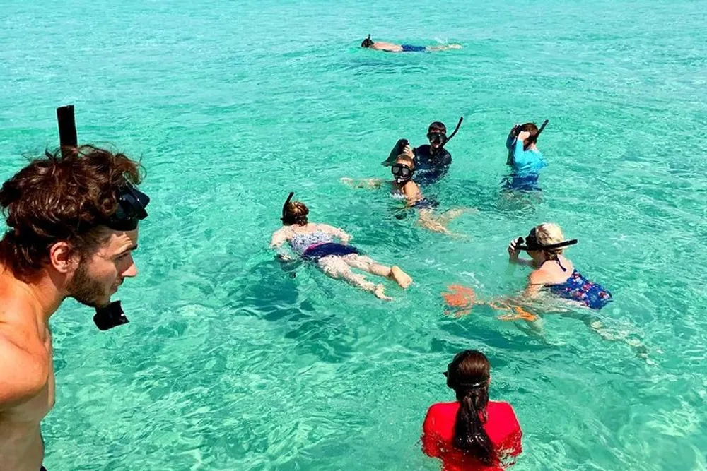 A group of people is enjoying snorkeling in clear turquoise waters with one person looking at the camera while others are already exploring underwater