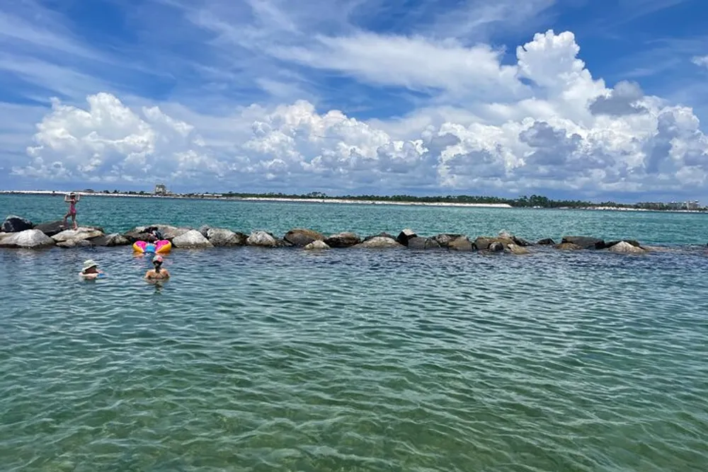 The image shows people enjoying clear shallow waters near a rock breakwater under a partly cloudy sky