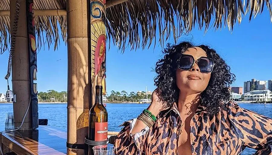 A person is striking a relaxed pose at a thatched-roof bar by the water, with a bottle of wine and tropical decor around.
