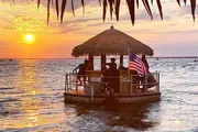 A small boat with a thatched roof bar and an American flag sails on calm waters during a beautiful sunset.