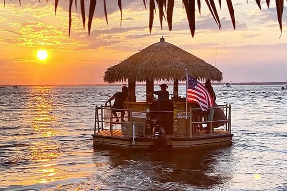 A small boat with a thatched roof bar and an American flag sails on calm waters during a beautiful sunset