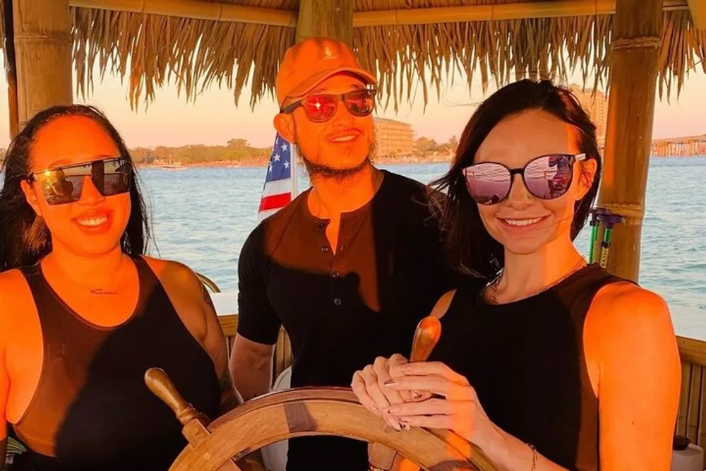 Three people are posing for a photo at the helm of a boat with a sunset and waterfront scenery in the background