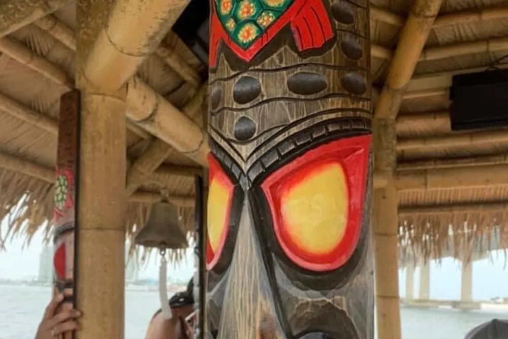 The image features a carved and colorfully painted wooden tiki pole inside a structure with a thatched roof and theres a blurred view of a person holding a cellphone in the background