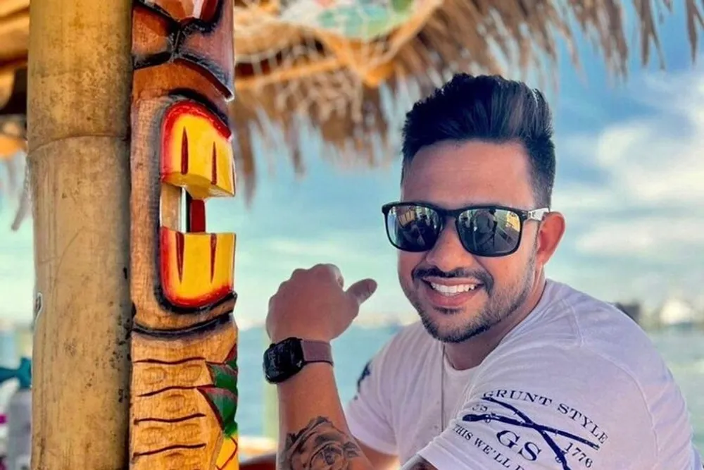 A smiling person with sunglasses is pointing at a colorful totem pole in a tropical setting