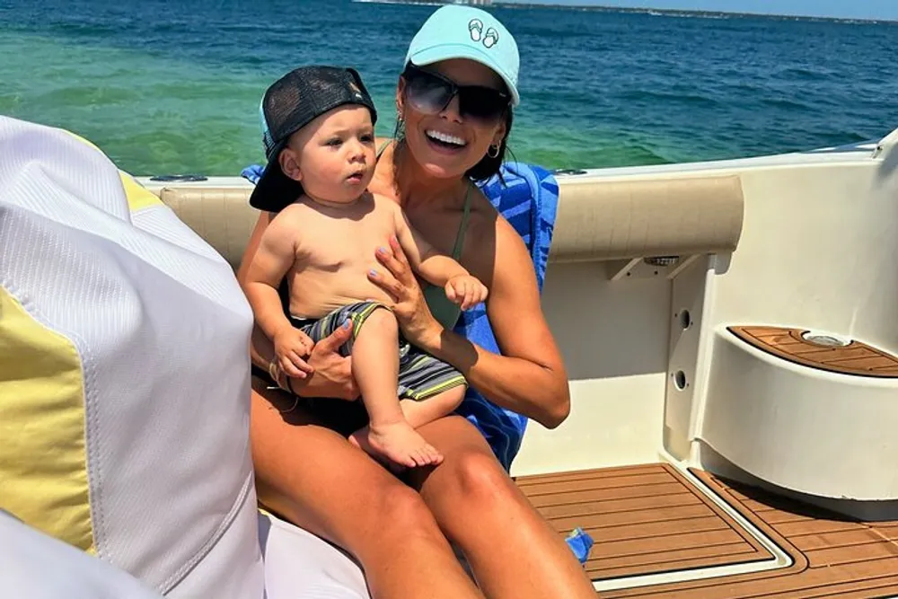 A child and a smiling woman wearing sunglasses and caps are enjoying time together on a boat with clear blue waters in the background