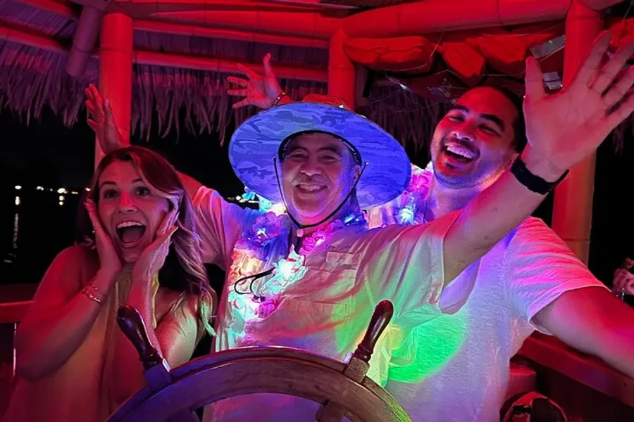 Three people are joyfully posing with hands up around a wooden ship's steering wheel under pink lighting, with one wearing a blue hat and lei, suggesting a festive or party atmosphere.