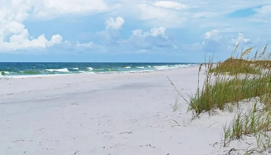 The image shows a serene beachscape with white sand, grass-topped dunes, and a calm sea under a partly cloudy sky.