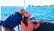 An older man is pointing something out to a woman by the water's edge on a boat with a view of the shoreline in the distance.