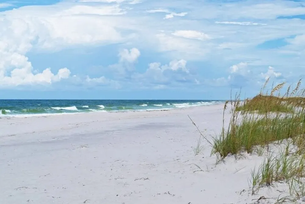 The image depicts a serene beach with white sand rolling waves a blue sky with scattered clouds and some grasses on the dunes