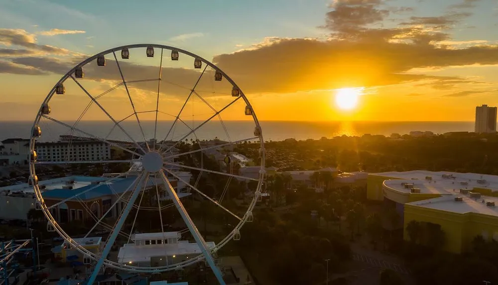 The image shows a towering Ferris wheel silhouetted against a vivid sunset sky overlooking a coastal area dotted with buildings