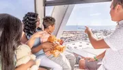 A family is enjoying a scenic view from inside a Ferris wheel gondola, with a sunny coastal cityscape stretching out in the background.