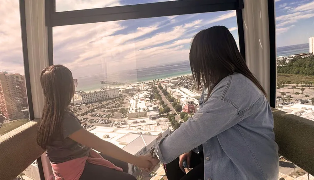 Two individuals are enjoying a scenic view from a high vantage point possibly within a Ferris wheel or observation tower overlooking a coastal cityscape and ocean horizon