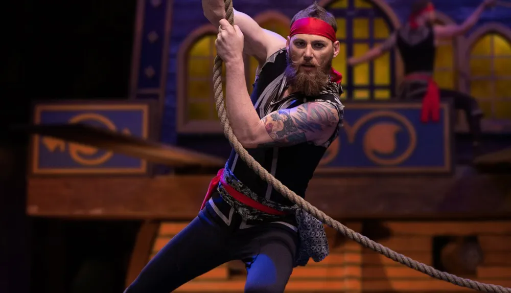 A bearded man dressed as a pirate swings on a rope in front of a ship-themed background