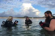 Three scuba divers are preparing for a dive in shallow water near the shore, under a sky with large cumulus clouds.