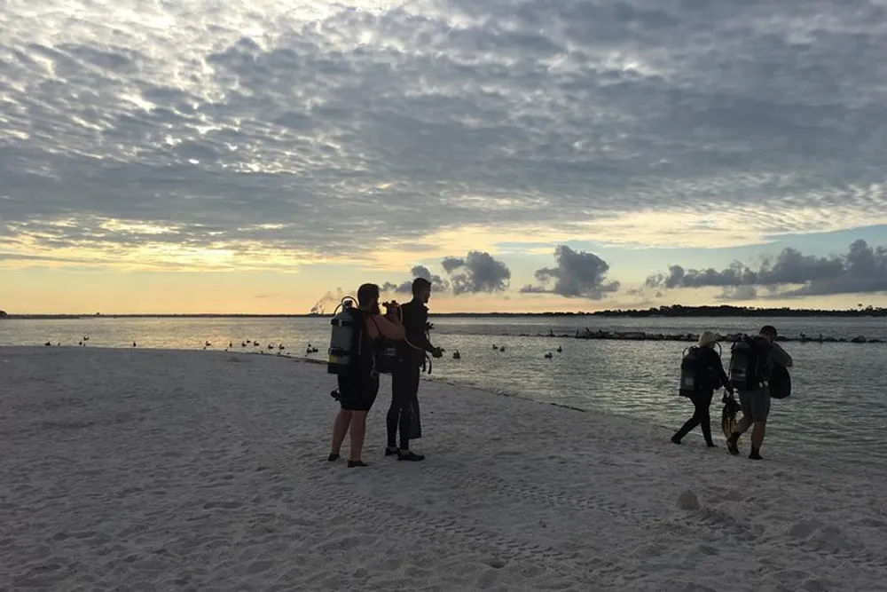 A group of people with scuba diving equipment is standing on a beach at dusk preparing for a dive