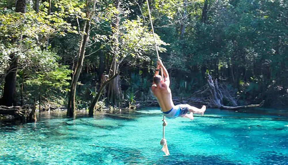 A person is joyfully swinging on a rope above a clear blue water body surrounded by lush greenery