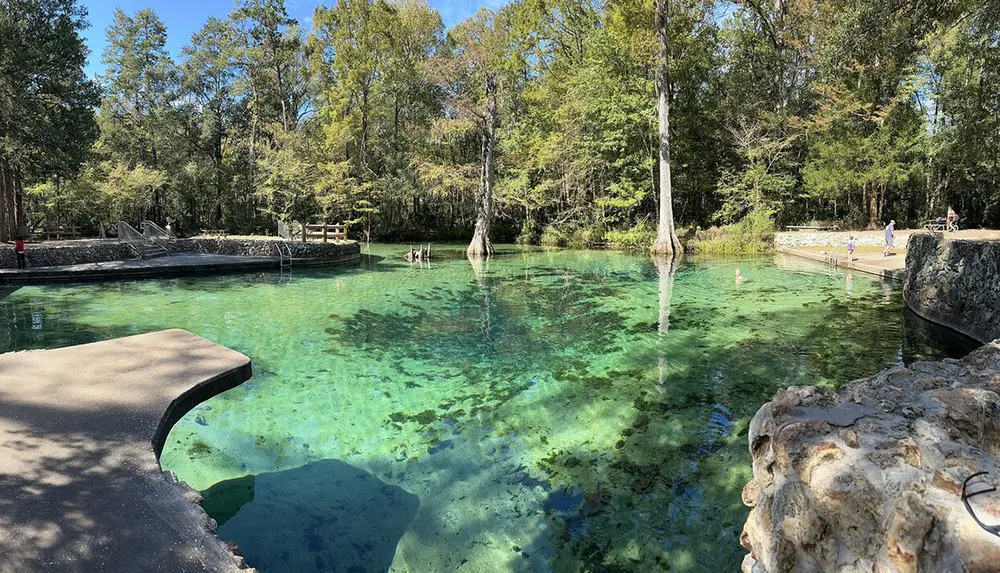 The image shows a serene crystal-clear natural spring surrounded by lush greenery with people enjoying the water and the natural setting