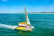 A colorful catamaran with passengers sails across blue waters near a coastline with some buildings in the distance.