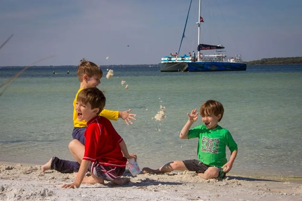 Three children are playing with sand on a sunny beach with a sailboat in the background